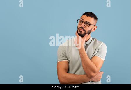 Pensive middle eastern guy thinking and touching chin, looking aside at free space, posing over blue background Stock Photo