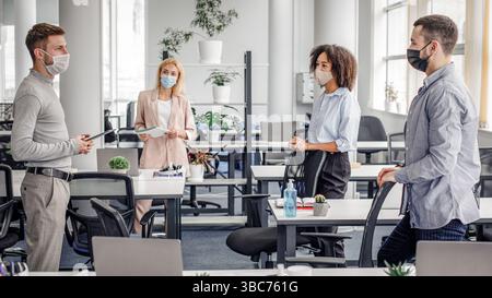 Urgent meeting of colleagues about creative ideas and business plans. Multiracial colleagues in protective masks maintain social distance and discuss Stock Photo