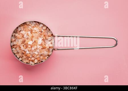 Coarse crystals of pink Himalayan salt in a metal measuring scoop against a pink background, top view Stock Photo