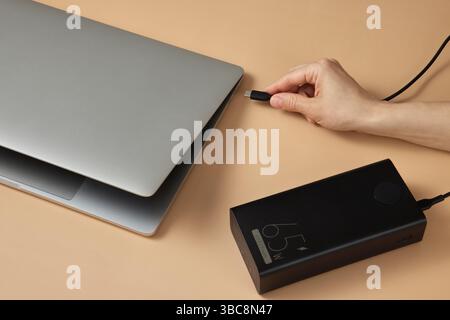 female hand connecting charger cable of power bank to laptop on beige background, close-up. Stock Photo