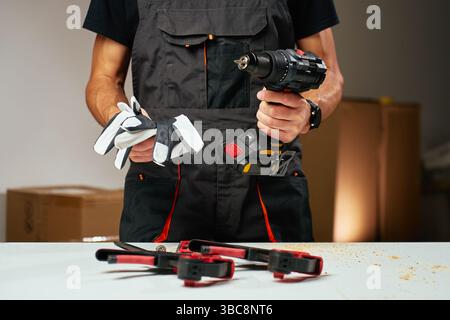 Person in work overalls holding cordless power drill and safety gloves at workbench with tools and wood shavings Stock Photo
