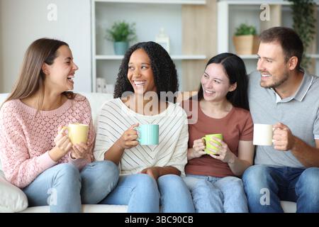 Four interracial friends talking sitting on a couch at home and drinking coffee Stock Photo