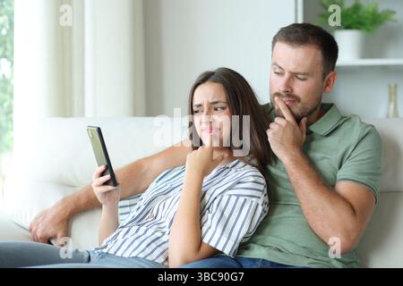 Suspicious couple checking phone content sitting in a couch at home Stock Photo
