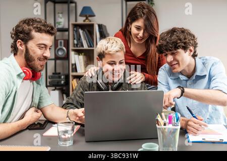 Smiling Gen-Z coworkers collaborating around a laptop, sharing ideas and planning a project in a bright, casual workspace; teamwork, startup and remot Stock Photo