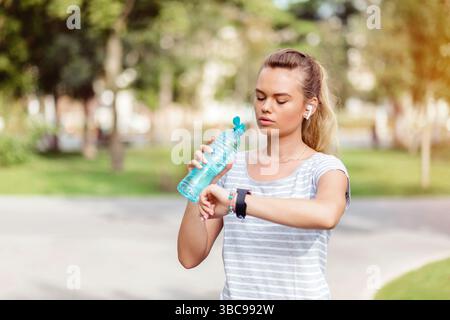 Young blonde woman jogging in park checking smartwatch and hydrating with water Stock Photo