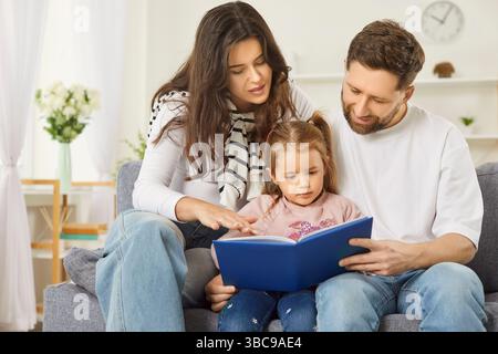 Close loving family at home, couple of parents teaching cute kid girl, read book, watching pictures Stock Photo