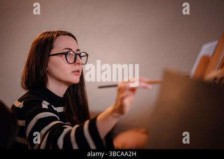Side view of young woman sketching on a canvas with pencil, fully focused during an indoor painting session. Stock Photo