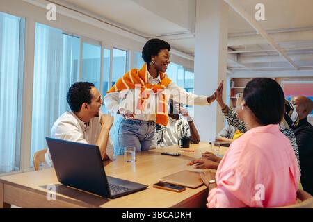 A group of diverse professionals celebrates a successful project in a modern agency office. A woman smiles and shares a high-five with a colleague, sy Stock Photo