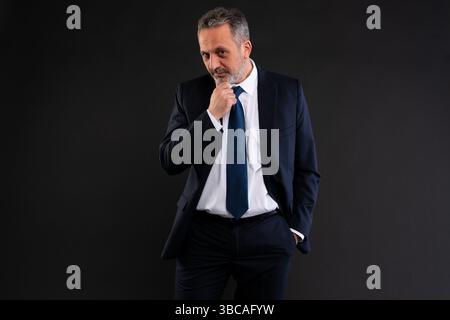 Confident mature businessman wearing a suit, looking thoughtful with hand on chin, against a dark background. Stock Photo