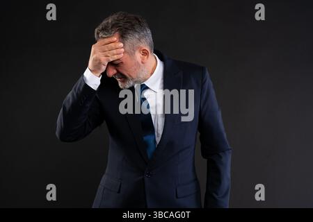 Mature businessman in dark suit touching his forehead in a gesture of frustration or regret, looking down, against a black background. Stock Photo