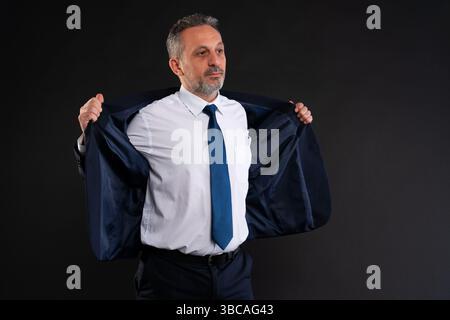 Confident mature man in shirt and tie putting on a dark blue suit jacket, preparing for work or business. Stock Photo