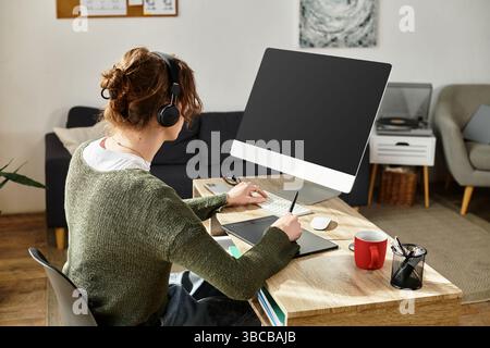 Focused young student wears headphones while taking notes at a desk in a cozy home office setting. Stock Photo