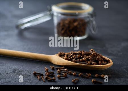 cloves spice in a wooden scoop and in glass storage jar on a black kitchen table, selective focus. Stock Photo