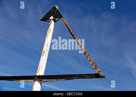 A damaged triangular nautical marker mounted on a weathered white pole stands against a vibrant blue sky, with its wooden structure showing signs of d Stock Photo