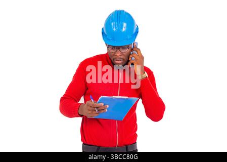 a young engineer wearing hard hat standing against white background looking at a notepad while communicating on the cell phone. Stock Photo