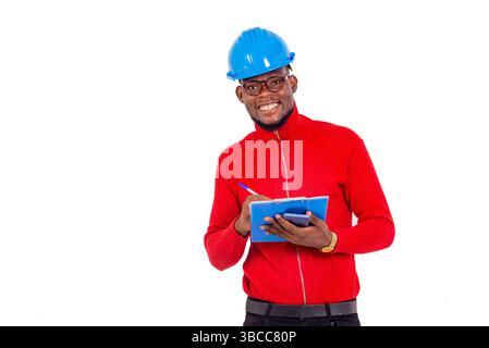a young engineer wearing a hard hat standing against white background writing on a notepad while looking at the camera smiling. Stock Photo