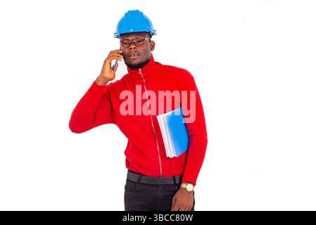a young engineer wearing safety helmet standing on white background talking on the cell phone while holding a notepad under the armpit. Stock Photo