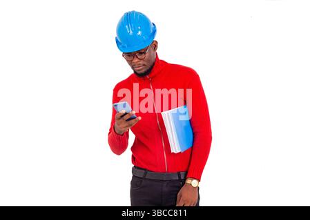 a young engineer wearing safety helmet standing on white background manipulating cellphone while holding notepad under armpit. Stock Photo