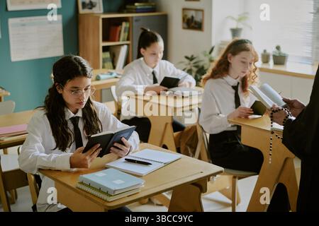 Group of high school students engaging in classroom activities while reading and studying together at desks. Teacher holding materials, supervising th Stock Photo