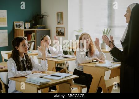 Engaged students raising hands while interacting with teacher in traditional classroom. Classroom features bookshelves and posters on the walls, evoki Stock Photo
