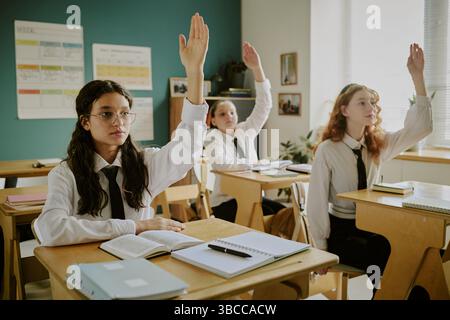 Group of students actively participating in classroom, raising hands to answer questions during lesson, books and notebooks on desks, sunlight illumin Stock Photo