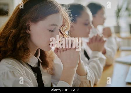 Schoolgirls in uniform holding hands and praying while sitting at desk in classroom, giving a sense of peace and unity, with concentration on their fa Stock Photo