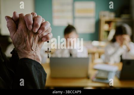 Elderly person holding rosary beads in focus with blurred background showing classroom setting, students engaged in study. Religious artifacts and lea Stock Photo