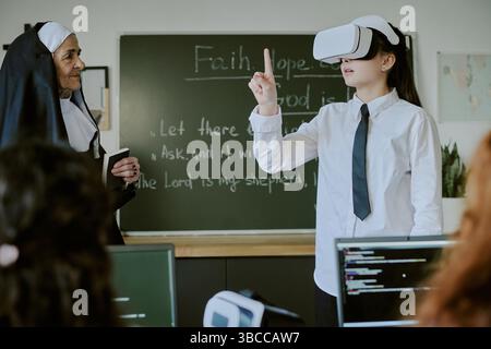 Schoolgirl using virtual reality headset in classroom engaging in educational activity with teacher observing nearby Stock Photo