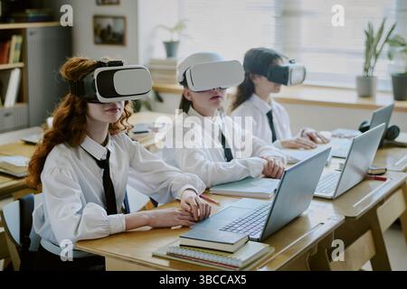 Students wearing virtual reality headsets and using laptops at classroom desk. Engaging in interactive learning experience with technology enhancing e Stock Photo