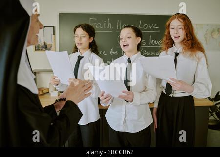 Group of students in uniform practicing singing while facing teacher in classroom with chalkboard backdrop. Teacher guiding students during rehearsal Stock Photo