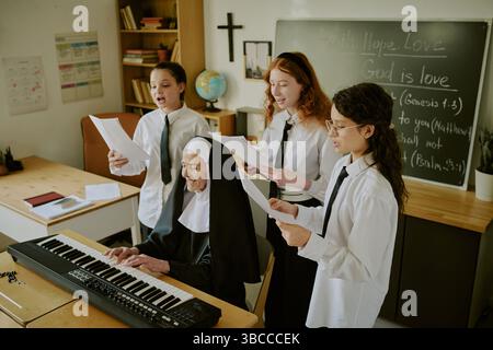 Elderly nun playing electric piano while students sing in classroom decorated with educational materials and cross on wall showing group engaged in mu Stock Photo