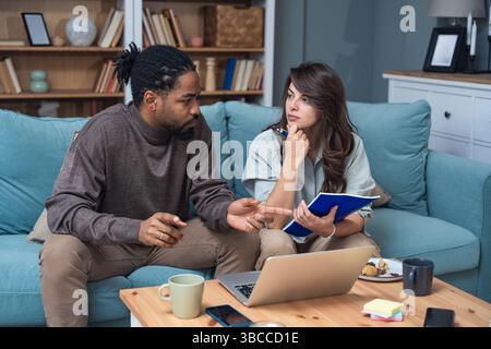 Young couple working remotely at home, analyzing documents and data, using laptop in cozy modern living room, discussing financial reports and busines Stock Photo