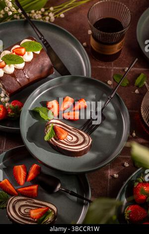 Chocolate swiss roll cake slices served on plates, garnished with fresh strawberries, mint leaves, and whipped cream, accompanied by a glass of coffee Stock Photo