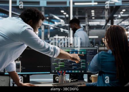 Data center supervisor overseeing admin using PC to visualize artificial intelligence neural networks node trees. Server room manager providing feedback to employee working to oversee AI systems Stock Photo