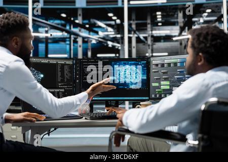 Man in wheelchair and coworker setting up data center gear needed to run artificial intelligence and machine learning. IT expert with disability and colleague in inclusive server room using AI tools Stock Photo