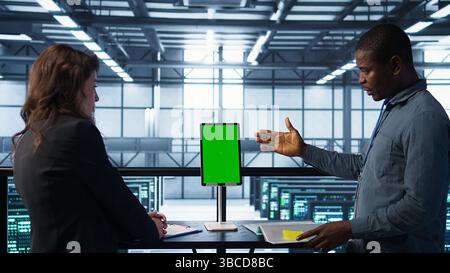Programmers in data center brainstorming, using mockup tablet, observing equipment analytics. Server hub IT coworkers use isolated screen device, adjusting system parameters, camera B Stock Photo