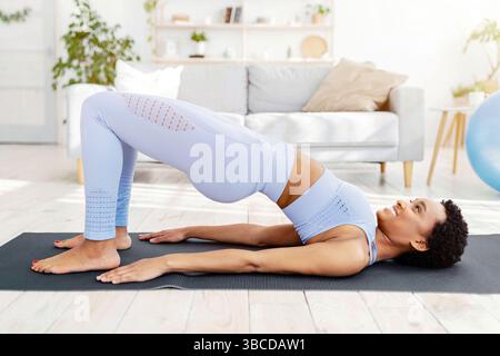 Sporty black woman doing exercises on yoga mat, standing in half bridge pose indoors, side view Stock Photo