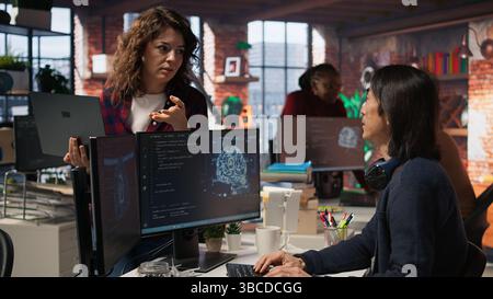 Startup intern holding laptop asks questions to senior technician coding advanced AI algorithm. Employee requests help from CTO manager in Silicon Valley workplace, camera A Stock Photo