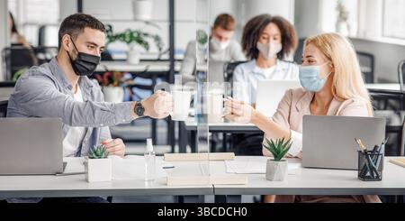 Coffee break in modern office and protecting health of workers. Young man and woman in protective masks clink cups through glass board in interior of Stock Photo