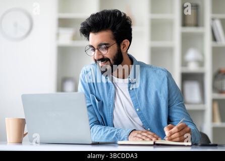 Online Lesson. Smiling Young Indian Man Using Laptop And Taking Notes Stock Photo