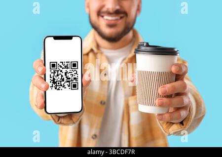 Young man holding smartphone with QR code on screen and coffee against blue background, closeup Stock Photo