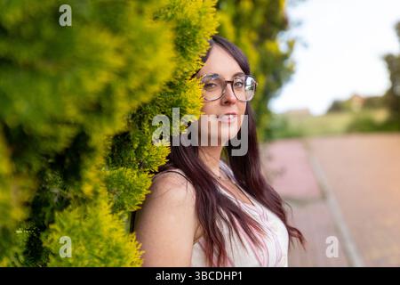 Young woman wearing glasses leaning against green bush in a park Stock Photo