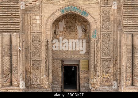 A pointed arch entrance at Magoki Attori Mosque in Bukhara, Uzbekistan, is framed by carved brick and faded blue tile with floral motifs. Stock Photo