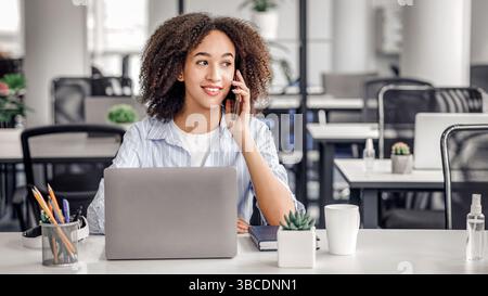 Working with modern technology and clients remotely. Friendly african american lady talking on phone with client, sitting at table with laptop in Stock Photo