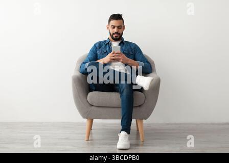 Young Arab guy using mobile phone, chatting online, sitting in armchair, surfing social media against white studio wall Stock Photo