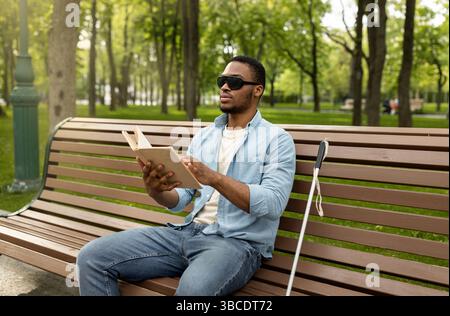 Young black visually impaired man sitting on bench in city park, reading Braille book outdoors Stock Photo