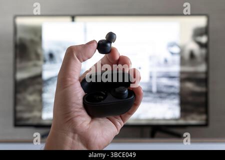 wireless headphones in the hands of a man on the background of a TV screen. watching TV with headphones at night. Stock Photo