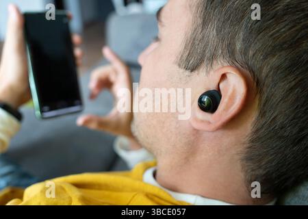 Close up of cheerful Caucasian adult man sitting on the sofa with wireless earphones in his ears and enjoying using modern smartphone Stock Photo