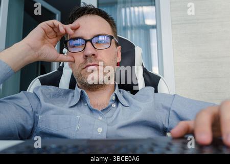 A young handsome man in a blue shirt is working at a computer on a light background. The programmer is focused on work. intellectual work Stock Photo
