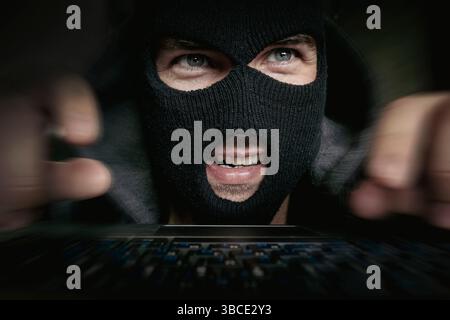 theft on laptop at night, internet crime online. Hacker in a balaclava standing in the darkness furtively stealing data of a laptop computer or insert Stock Photo